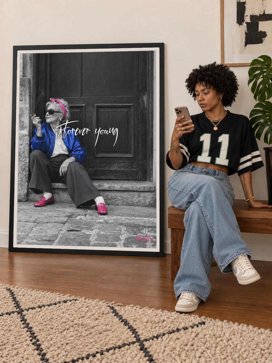 Forever Young print, 36 × 48 inch, large black frame beside a seated woman in a cropped black jersey and wide jeans checking her phone